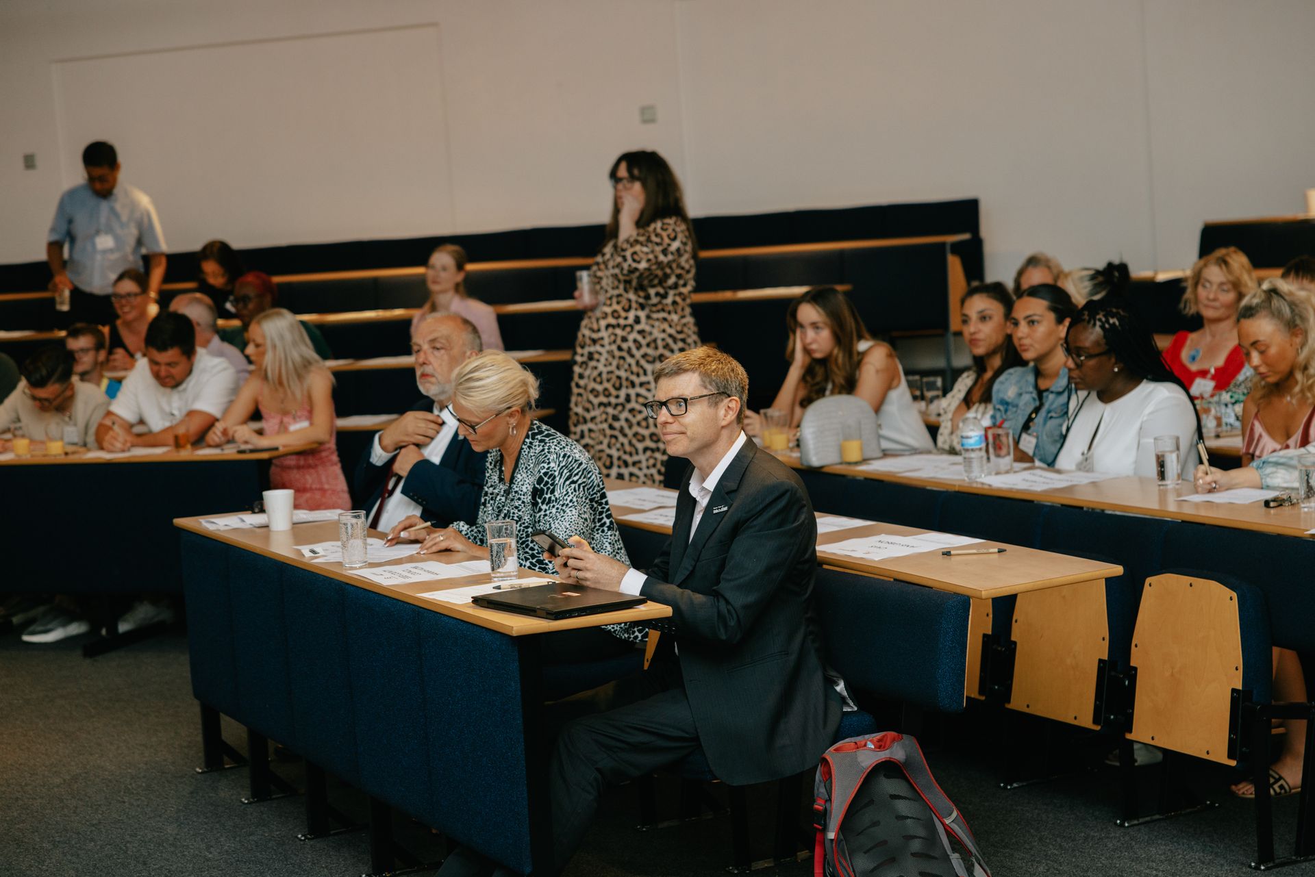 A group of people are sitting at long tables in a lecture hall.