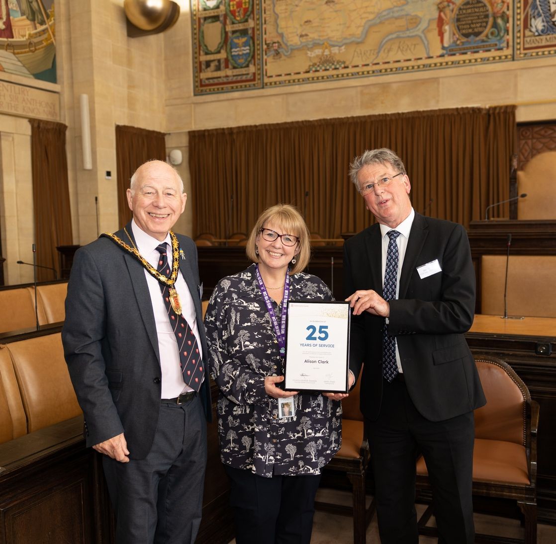 Three people are standing in a room holding a certificate that says 25 on it.