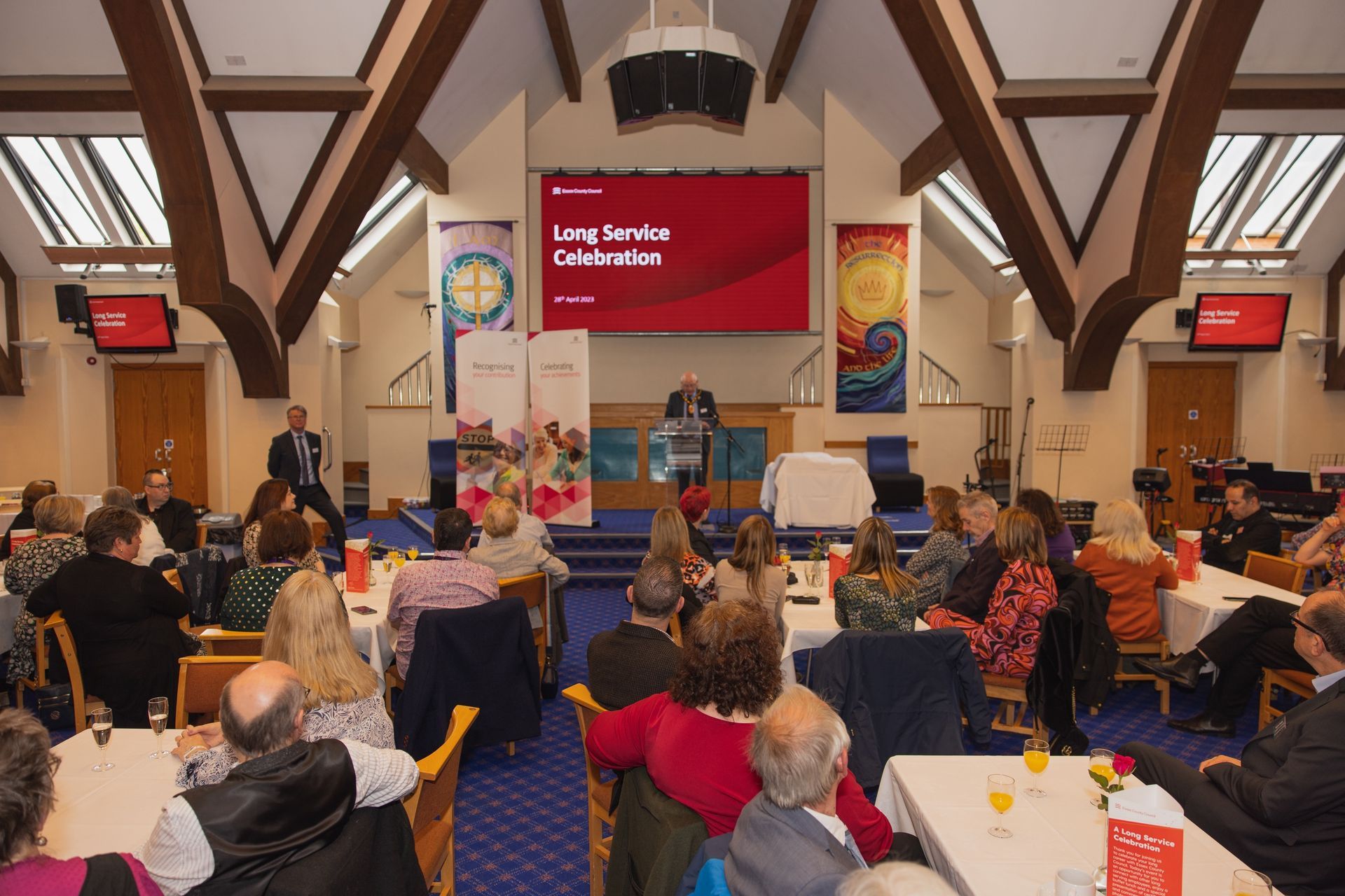 A large group of people are sitting at tables in a church watching a presentation.