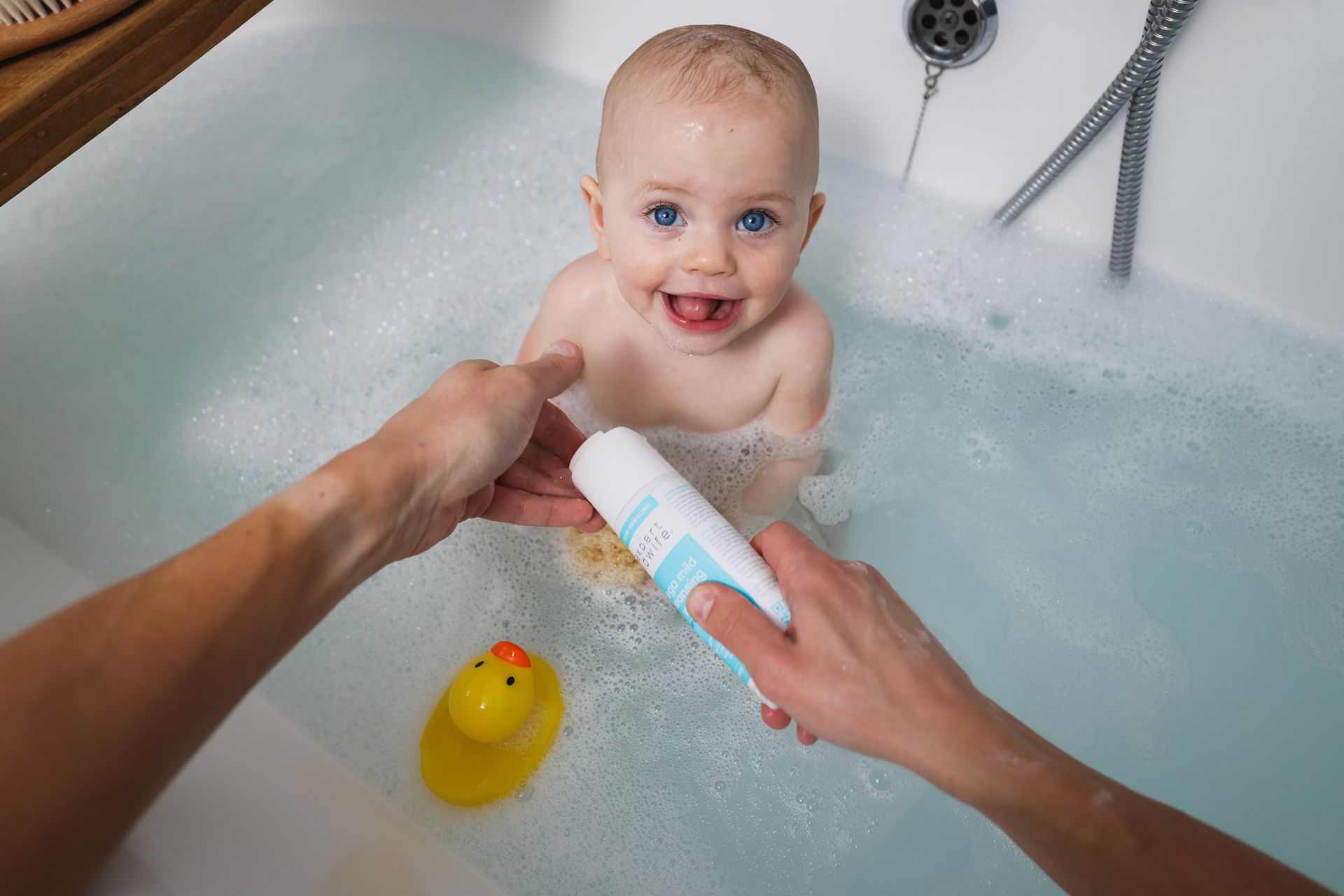 A baby is taking a bath in a bathtub while a person holds a bottle of shampoo.
