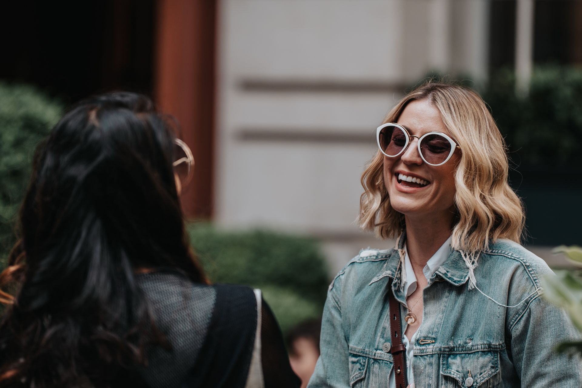 A woman wearing sunglasses is smiling while talking to another woman.