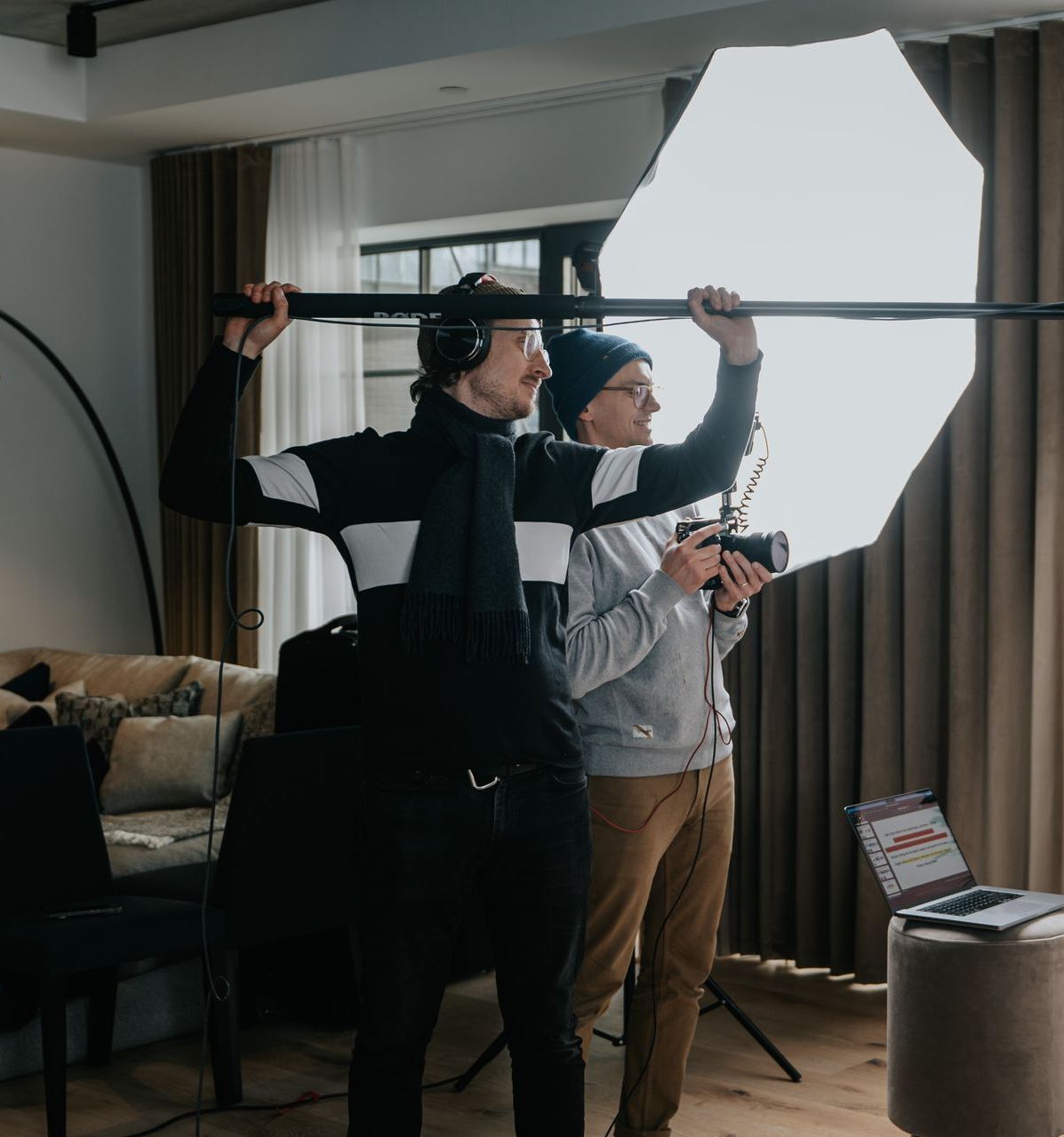 Two men are standing in a living room holding umbrellas and cameras.