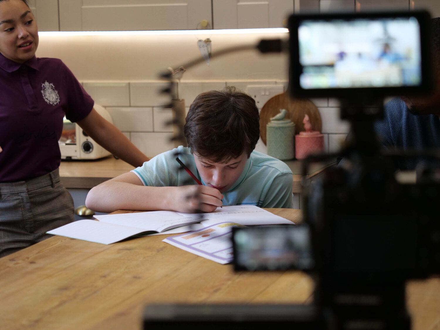 A woman is standing next to a boy who is writing on a piece of paper.