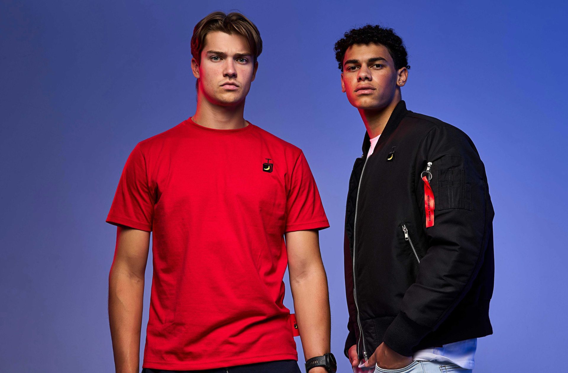 Two young men are standing next to each other wearing red shirts and black jackets.