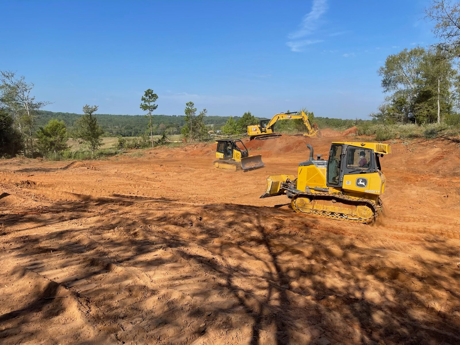 Two yellow excavators on a dirt clearing under a blue sky, with trees in the background.