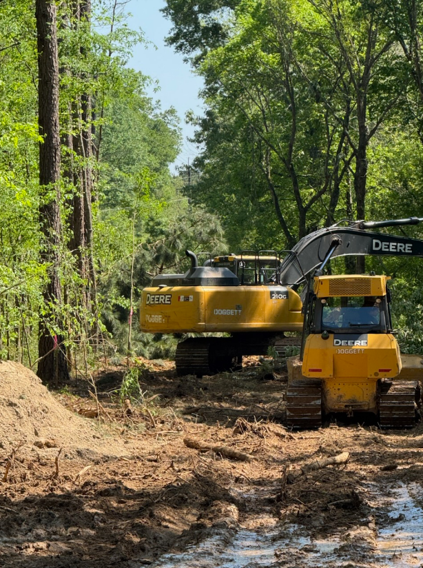 Yellow excavator and compact track loader on a muddy forest trail, clearing land among tall trees
