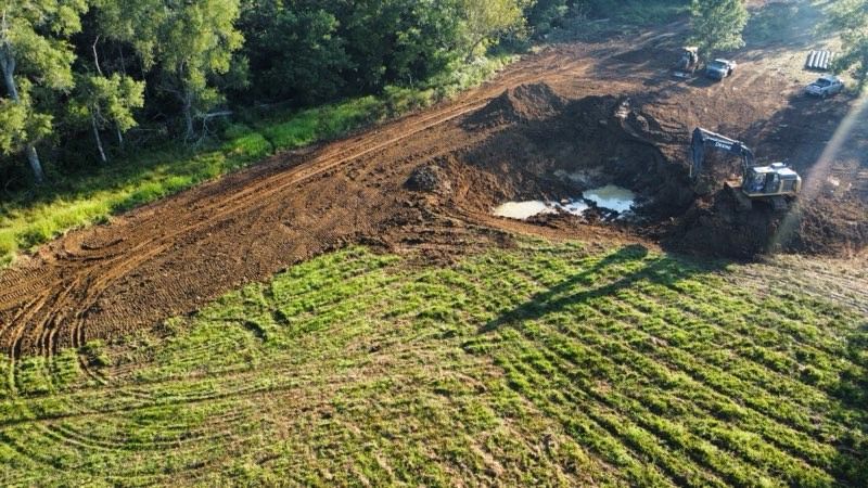 Mudslide in a rural field beside a road, with excavator clearing debris near a wooded area.