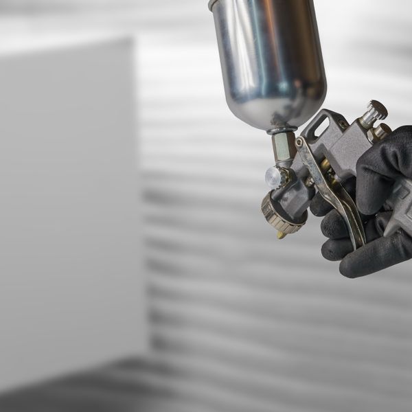 Gloved hand holding a spray paint gun aimed at a white wall in a workshop