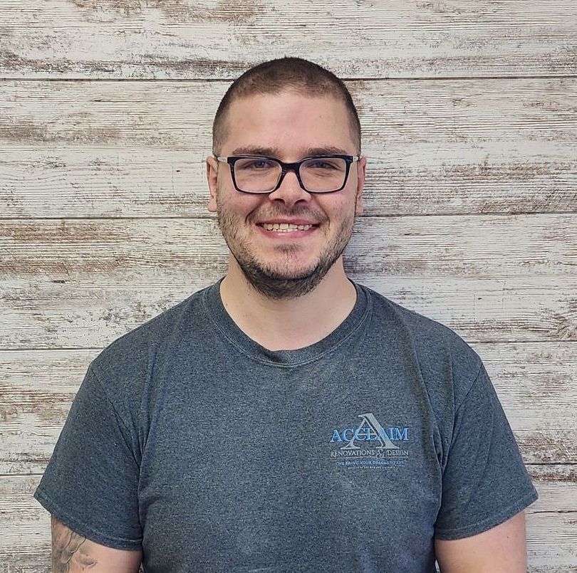Man wearing glasses and a gray shirt smiles in front of a wood-paneled wall.