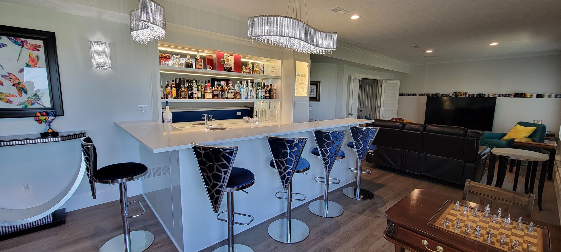 A home bar with stools, lit shelves, and a chess table, in a room with a seating area.