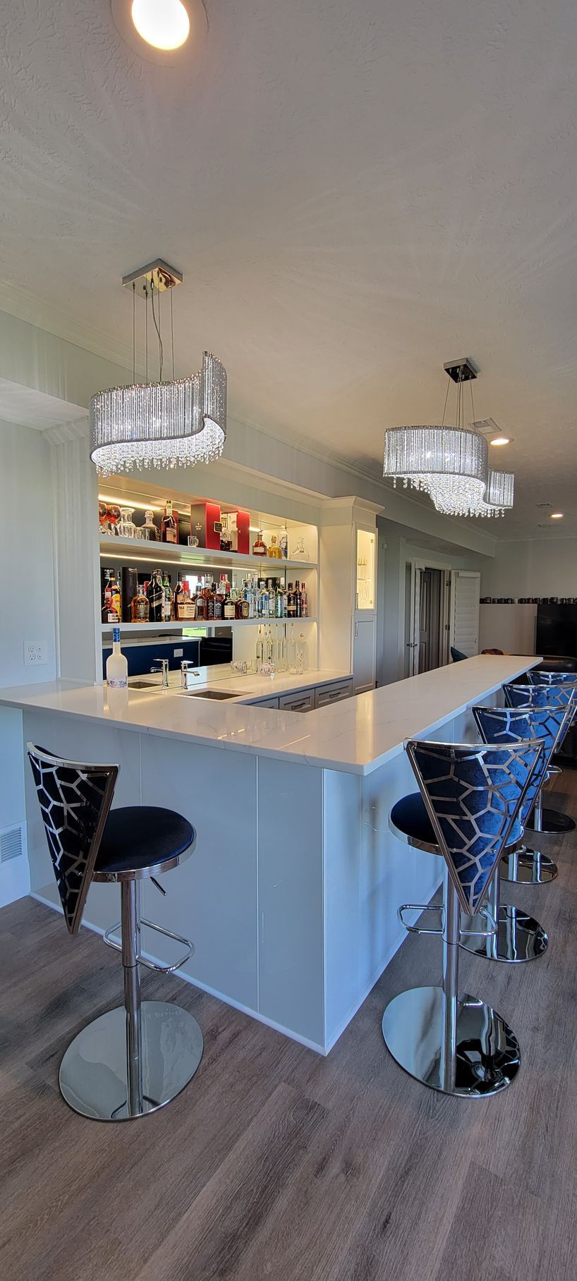 A home bar with white counter, liquor shelves, pendant lights, and bar stools.