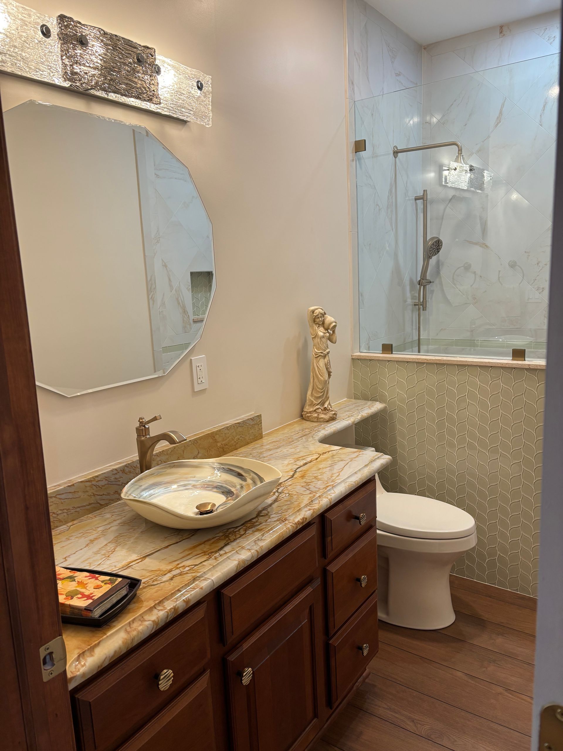 Bathroom with brown cabinets, gold countertop, round mirror, and a glass shower.
