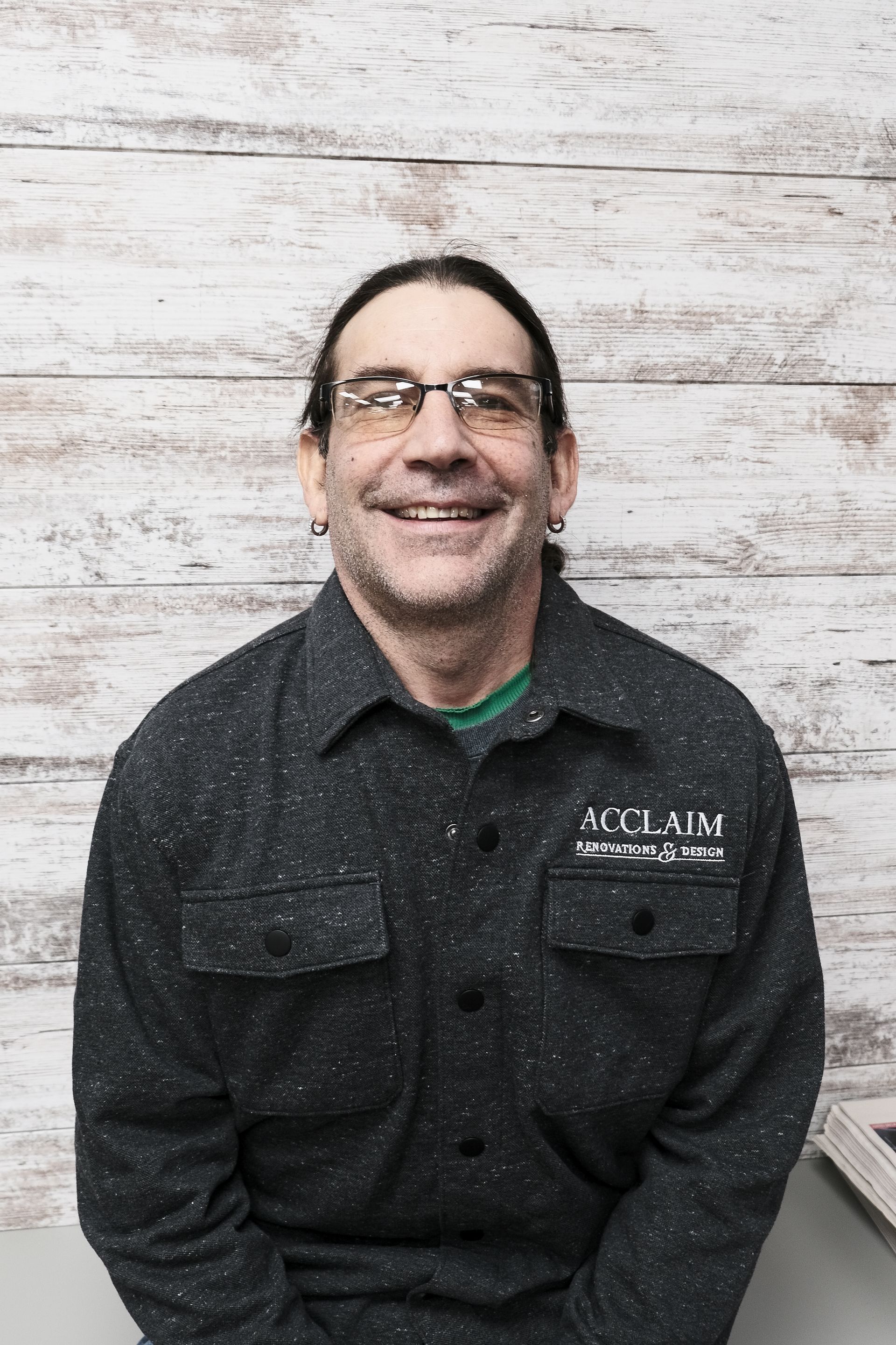 Man with glasses and long hair smiles in a dark flecked shirt, in front of a wood-paneled wall.