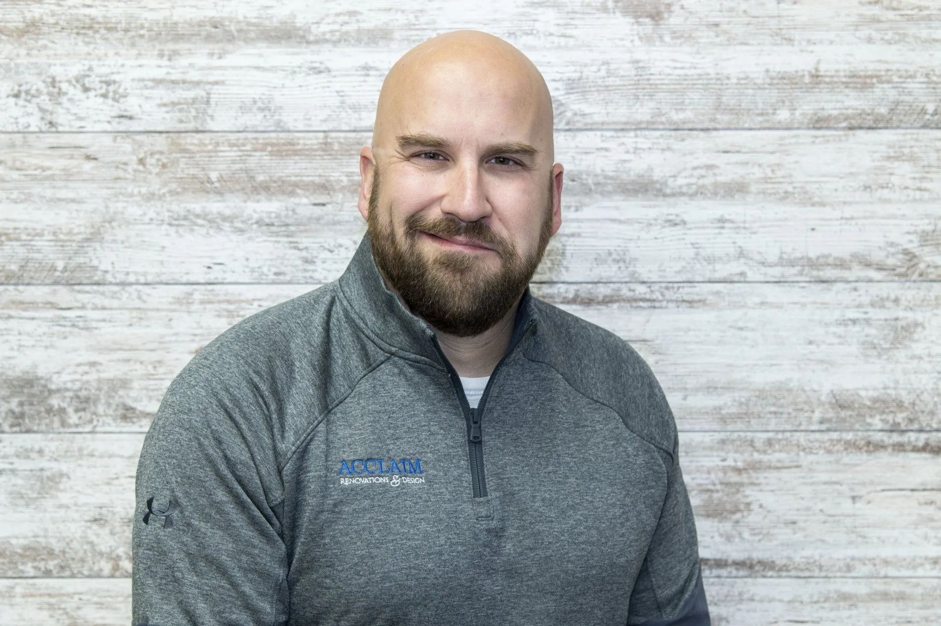 Bald man with beard smiles, wearing a gray pullover. Light wood-paneled background.