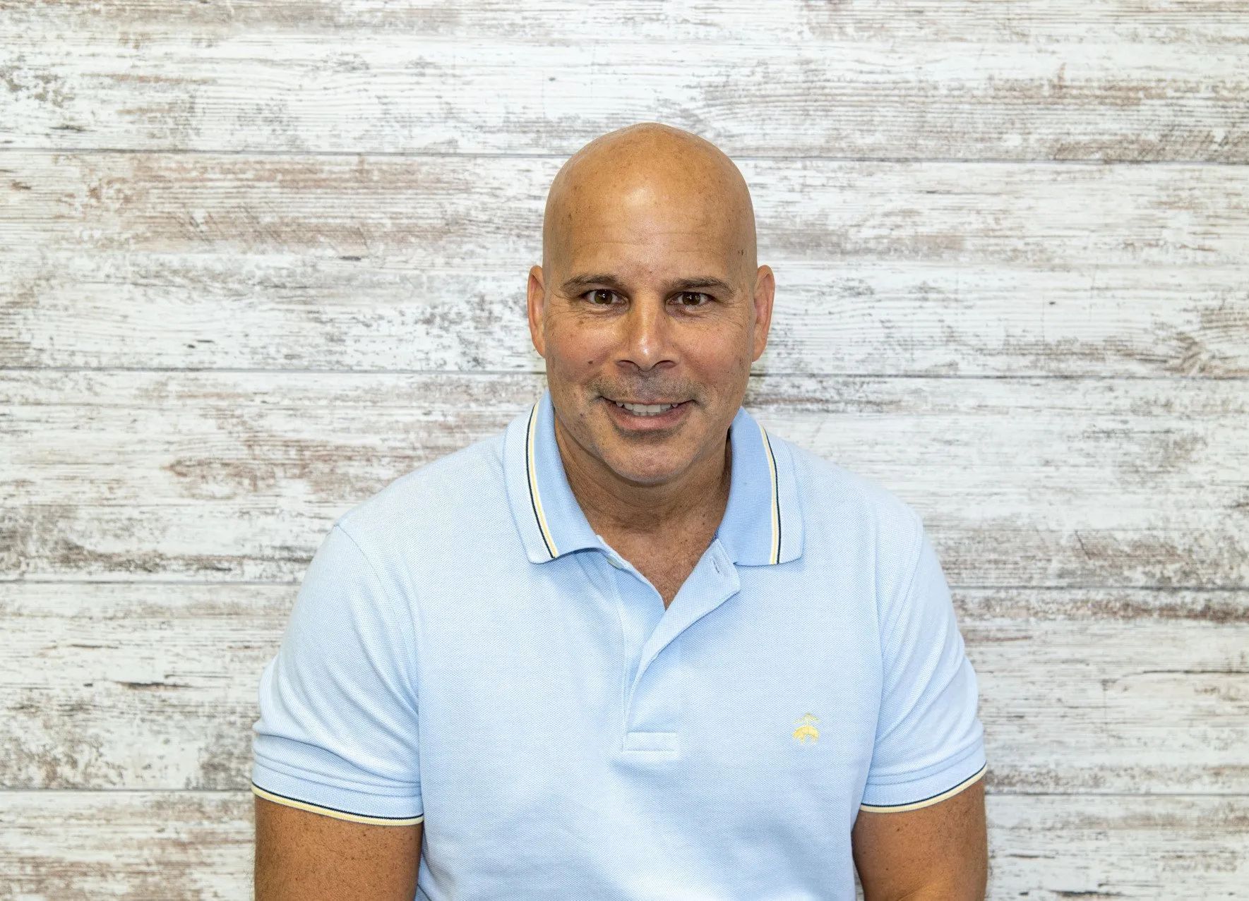 Bald man in light blue polo shirt smiles in front of a white wood-paneled wall.