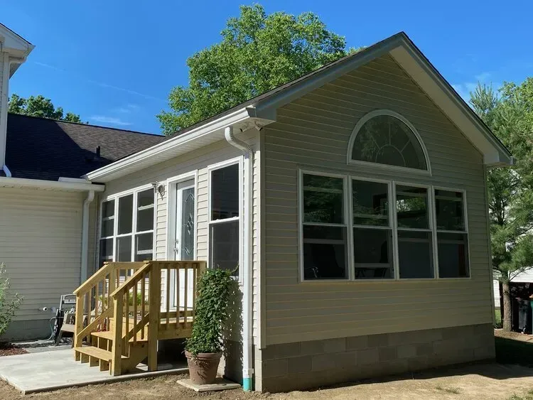 A sunroom addition to a house with windows, a door, and a small deck; tan siding and a blue sky.