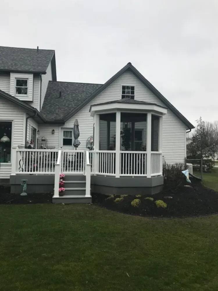 White house with deck and screened porch, surrounded by lawn and flower beds under a gray sky.