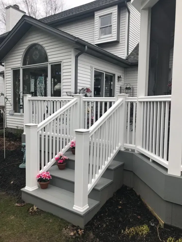 White deck with stairs, railings, and potted pink flowers leading to a gray house with windows.