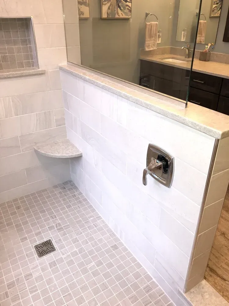 Modern shower with white tiled walls, glass partition, built-in seat, and dark vanity visible in background.