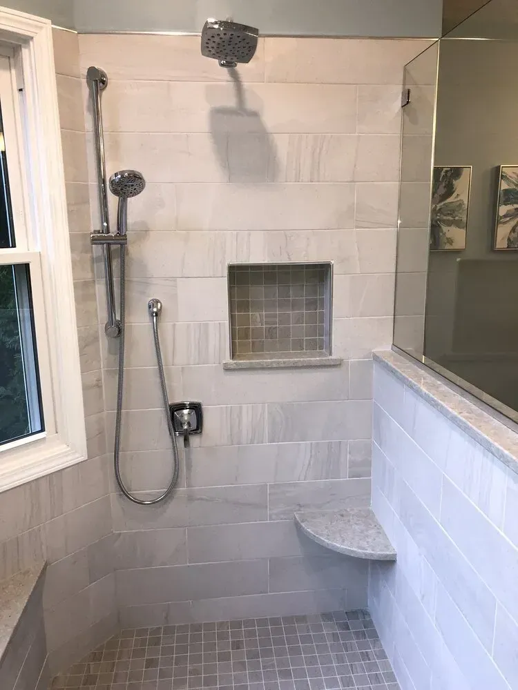 Shower with gray rectangular tiles, a built-in seat, recessed shelf, and handheld showerhead.