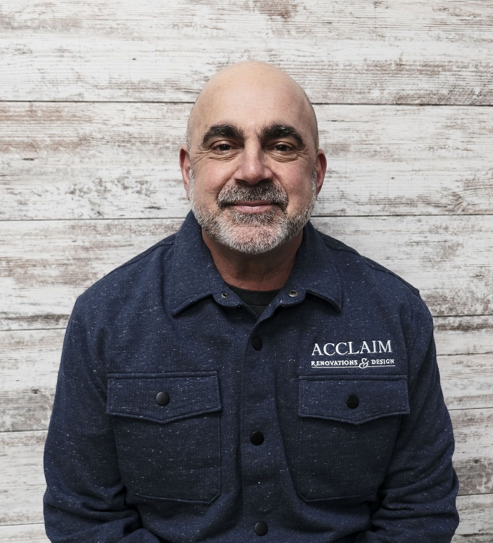 Bald man with a graying beard, wearing a blue button-up shirt, standing in front of a wood background.