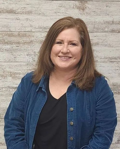 Woman smiling, wearing a blue jacket over a black shirt, in front of a wood-paneled wall.