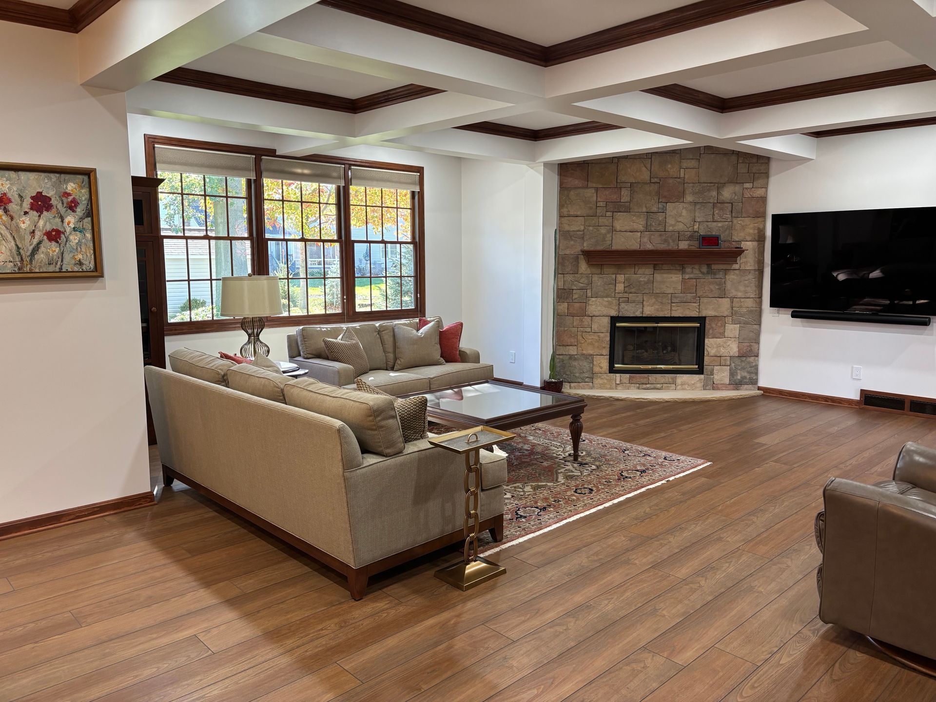 Living room with stone fireplace, brown wood floor, light beige sofas, and a coffered ceiling.