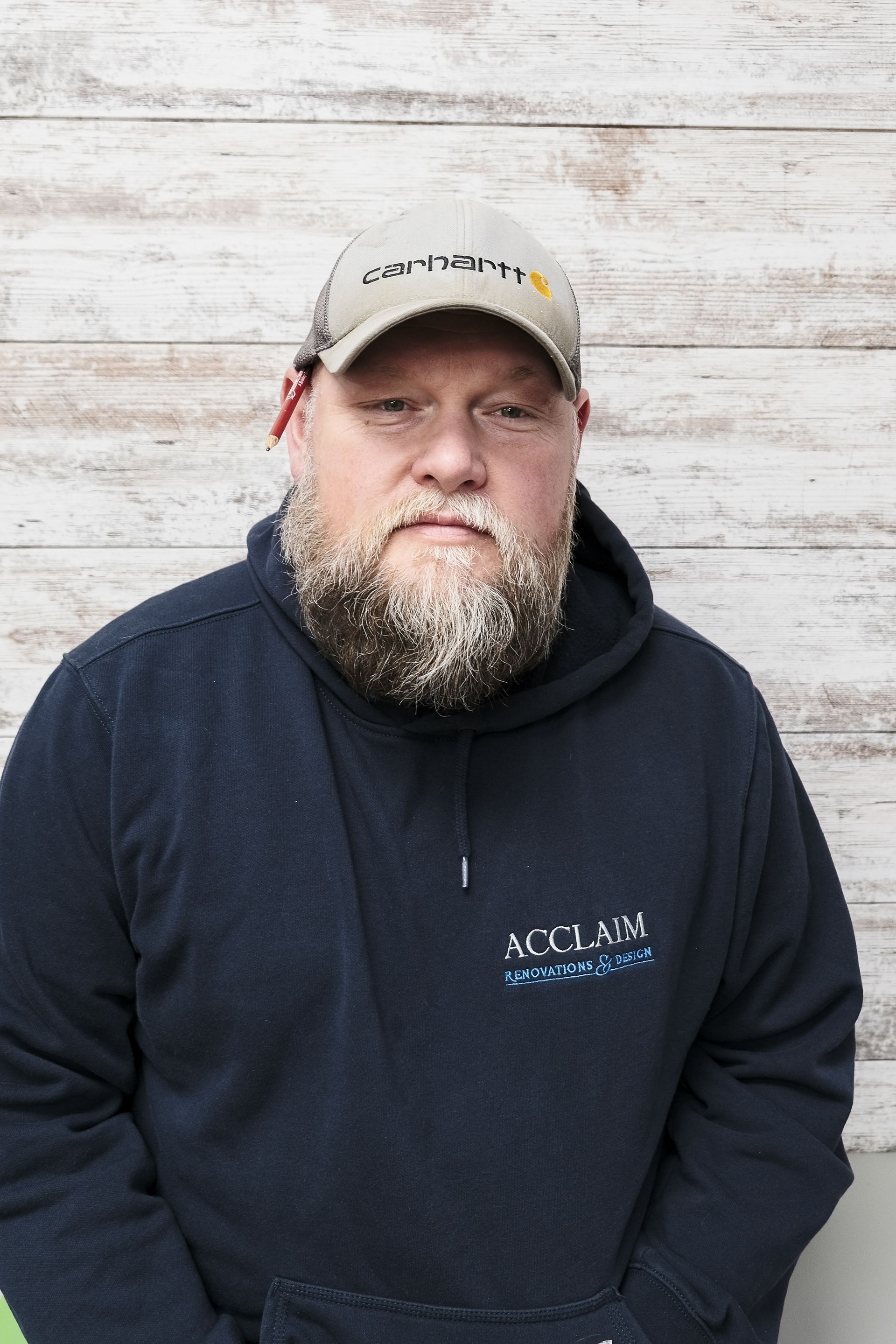 Man wearing a Carhartt cap and a blue hoodie stands in front of a wood-paneled wall, looking at the camera.