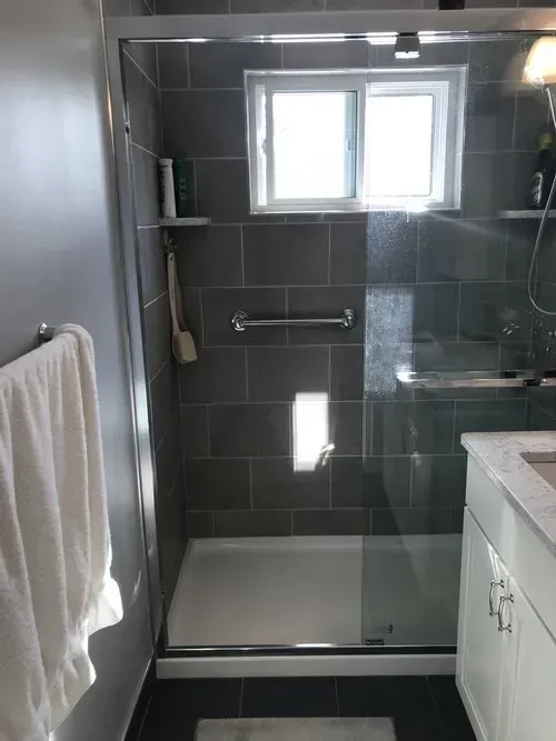 Bathroom with gray tiled shower, grab bar, glass door, window, and white vanity.