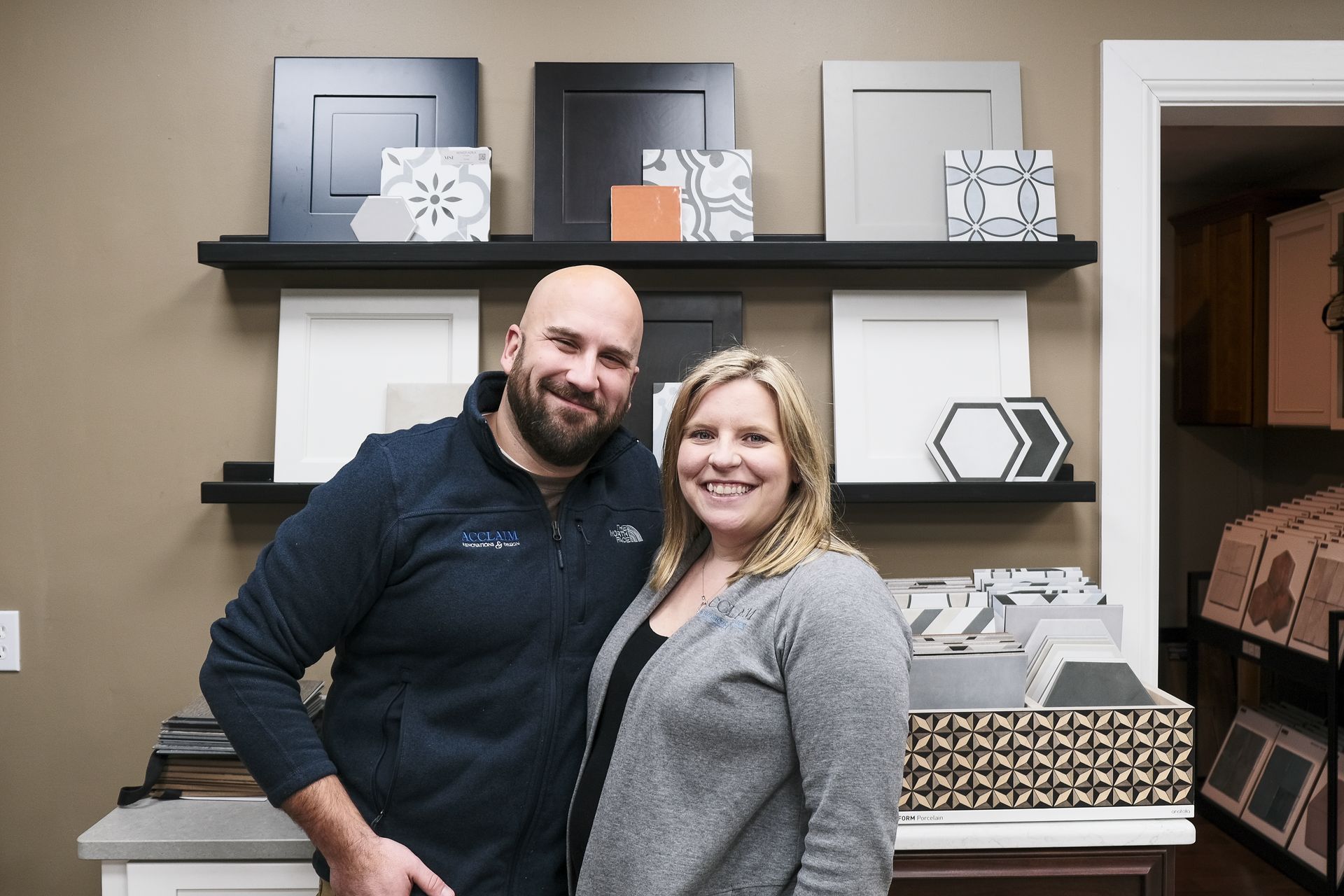 Couple smiling, standing in front of shelves displaying cabinet doors and tile samples in a showroom.