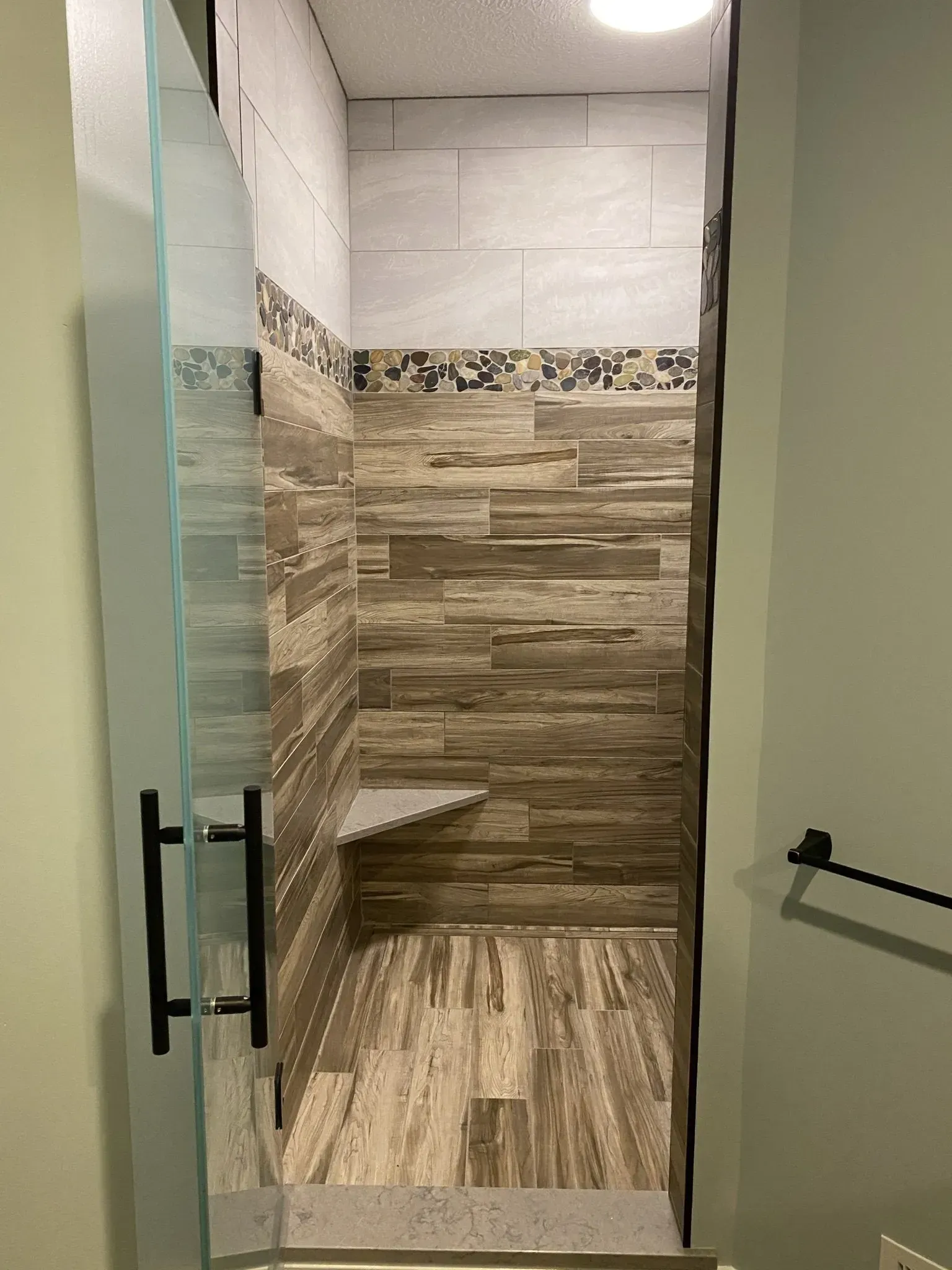 Shower with glass door, wood-look tile, mosaic border, and corner seat.