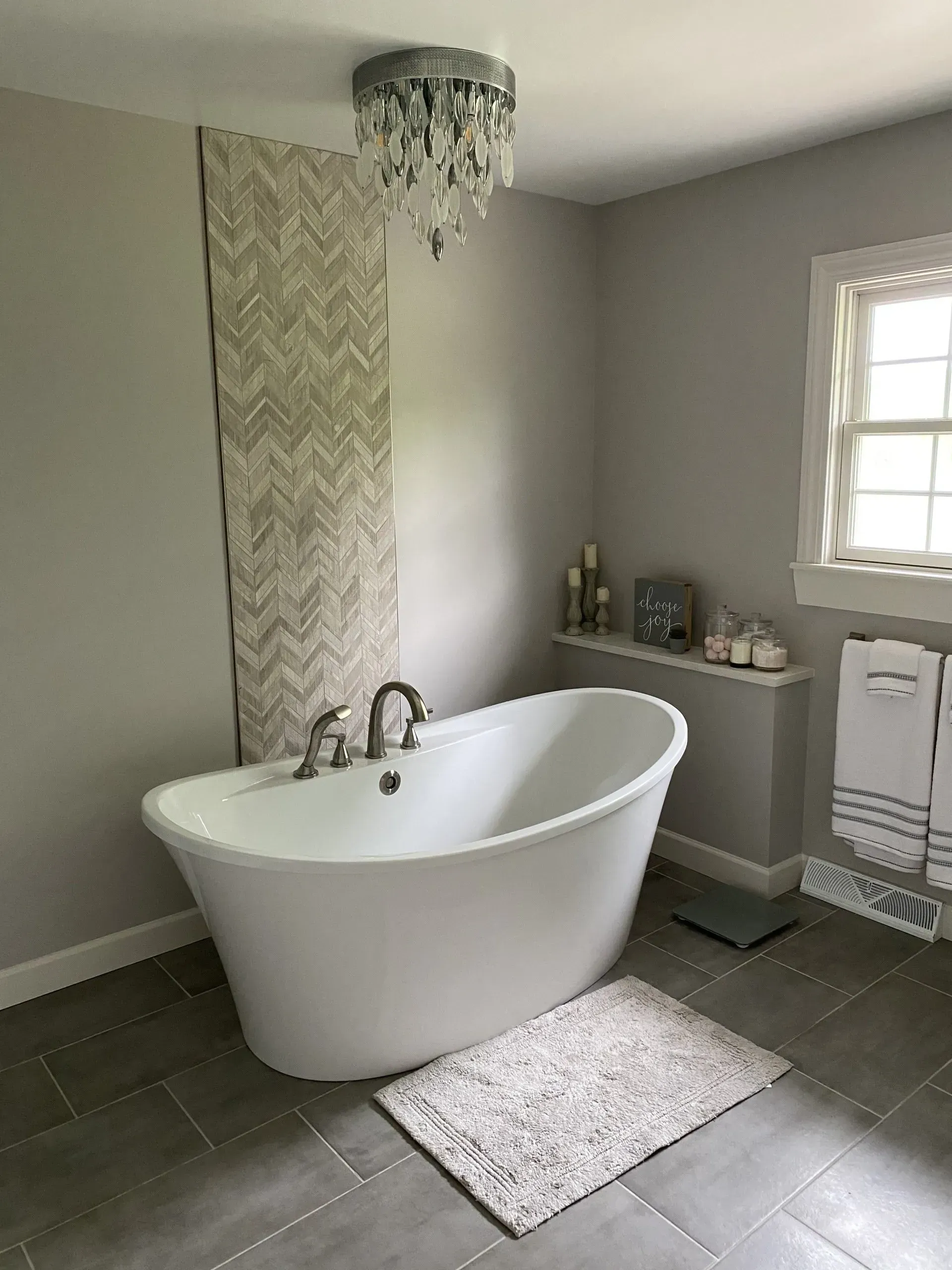 Elegant white soaking tub in a gray-walled bathroom, with a patterned tile accent, and a decorative chandelier.