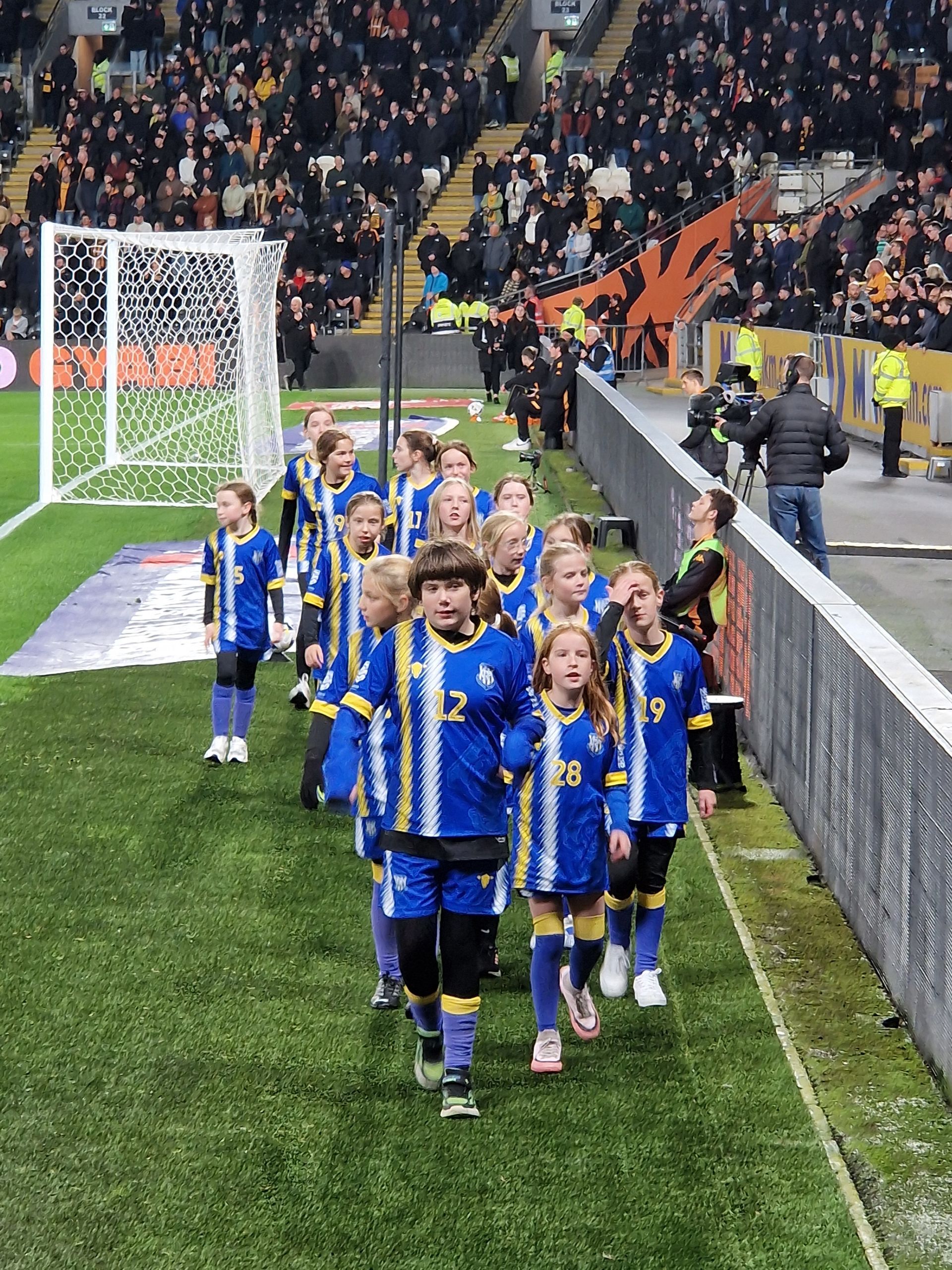 Children in blue and yellow soccer uniforms walk onto a green field. A crowd watches in the background.