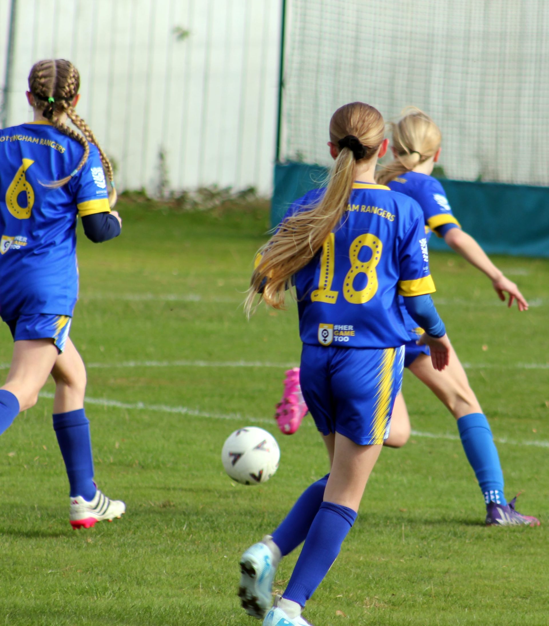Girls in blue soccer uniforms playing soccer on a green field. Number 18 kicks the ball.