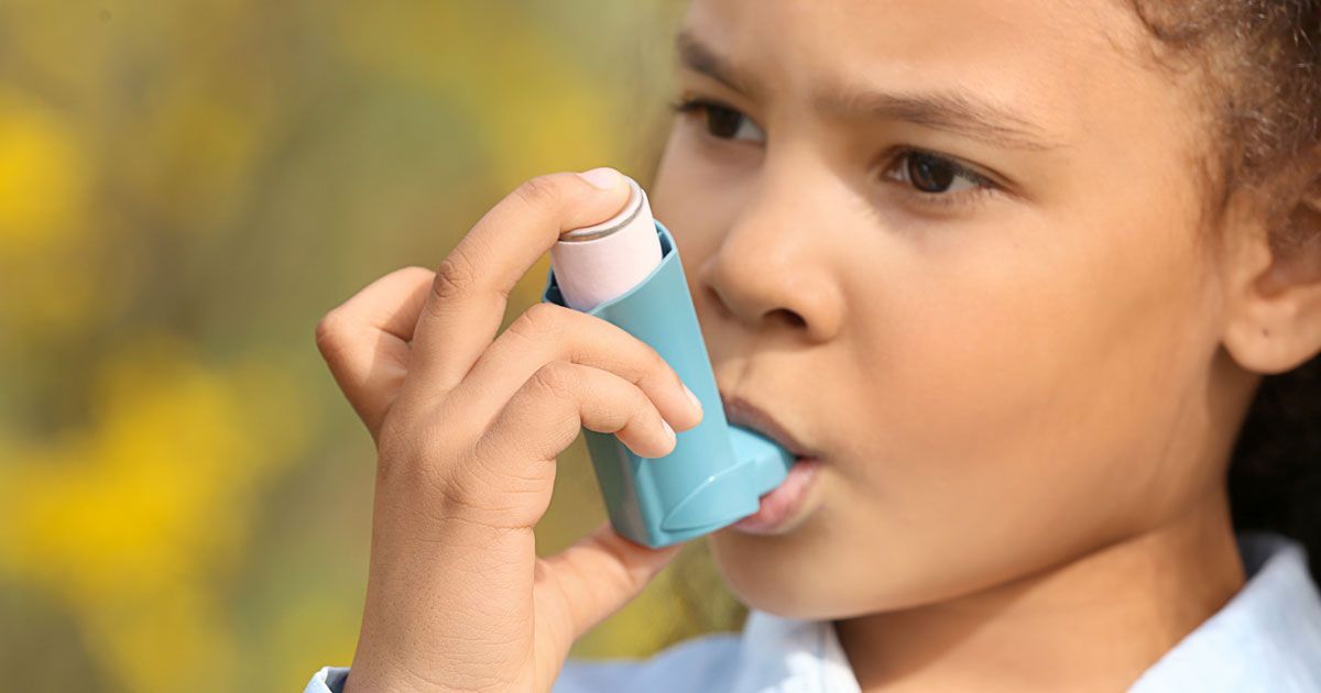 Girl using an inhaler outdoors, holding it to her mouth, light blue device.