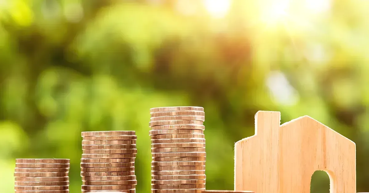 Stacks of coins beside a small wooden house on a blurred green background