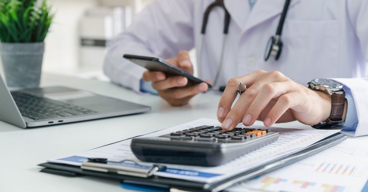 Doctor using calculator at desk with laptop and clipboard during office work
