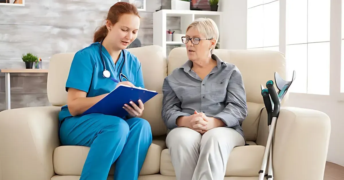 Caregiver in blue scrubs speaking with a seated patient in a bright living room