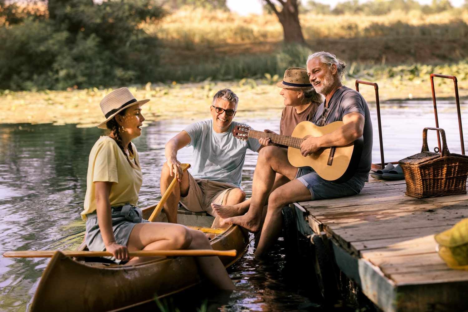 Three people relaxing on a dock by a river at sunset, with a guitar and canoe nearby.