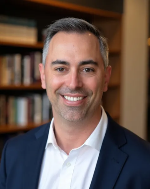 Smiling man in a navy suit and white shirt, standing in front of a blurred bookshelf background