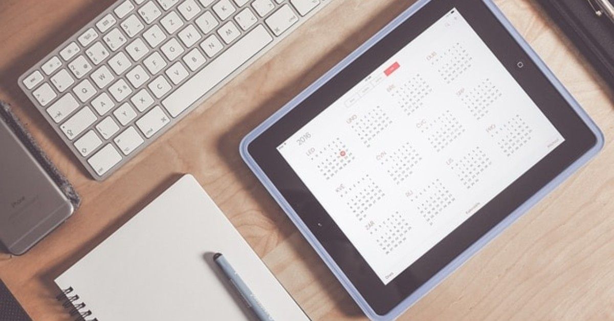 Desk with keyboard, notebook, and stylus beside a tablet displaying a calendar or schedule.