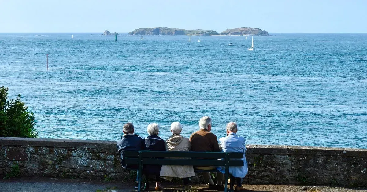 Four people sit on a bench by the sea, looking toward a calm blue horizon and distant islands.