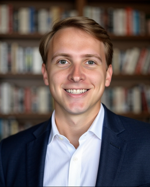 Headshot of a smiling person in a navy blazer and white shirt against a gray background