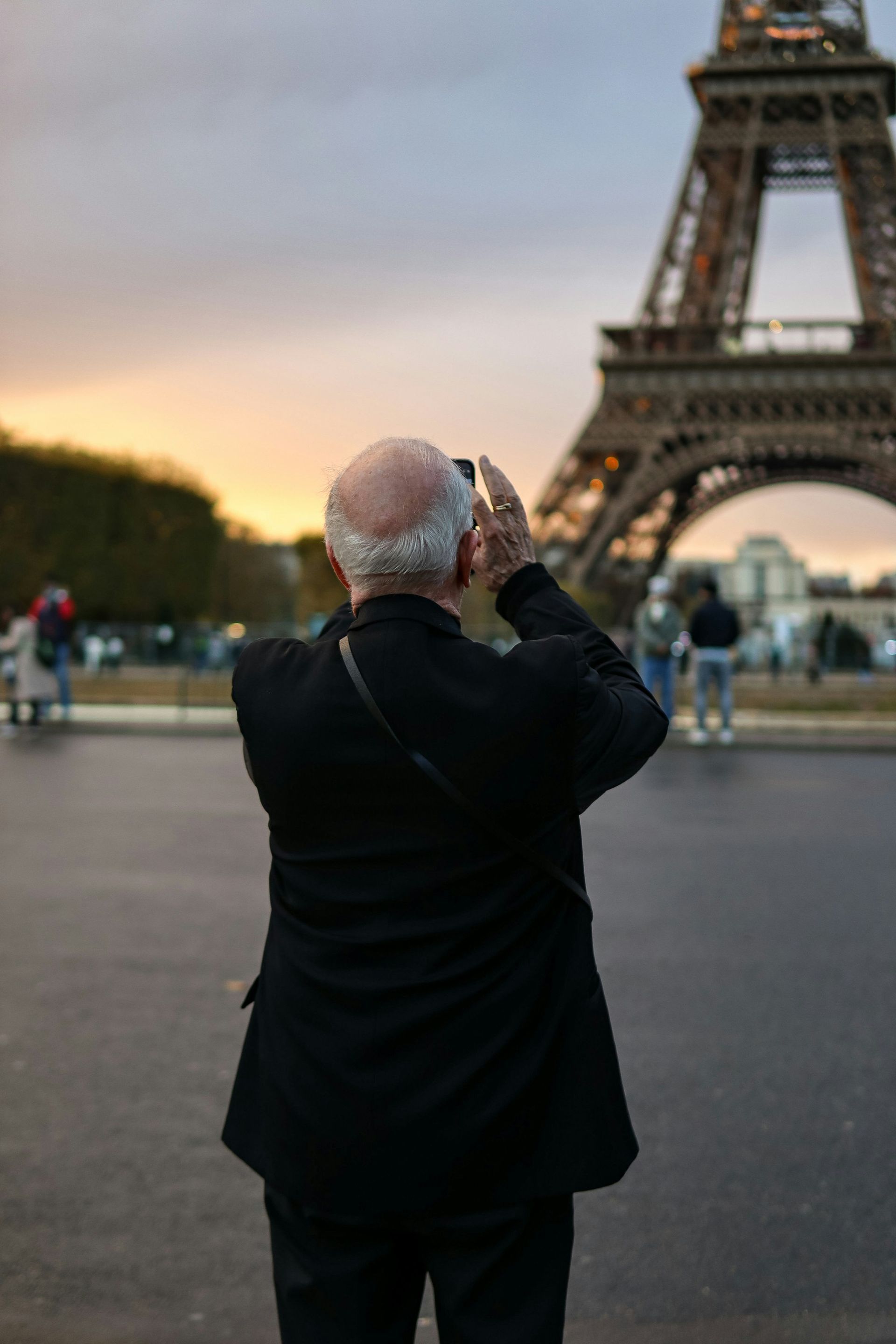 Person photographing the Eiffel Tower at dusk, with city lights and a sunset sky in the background