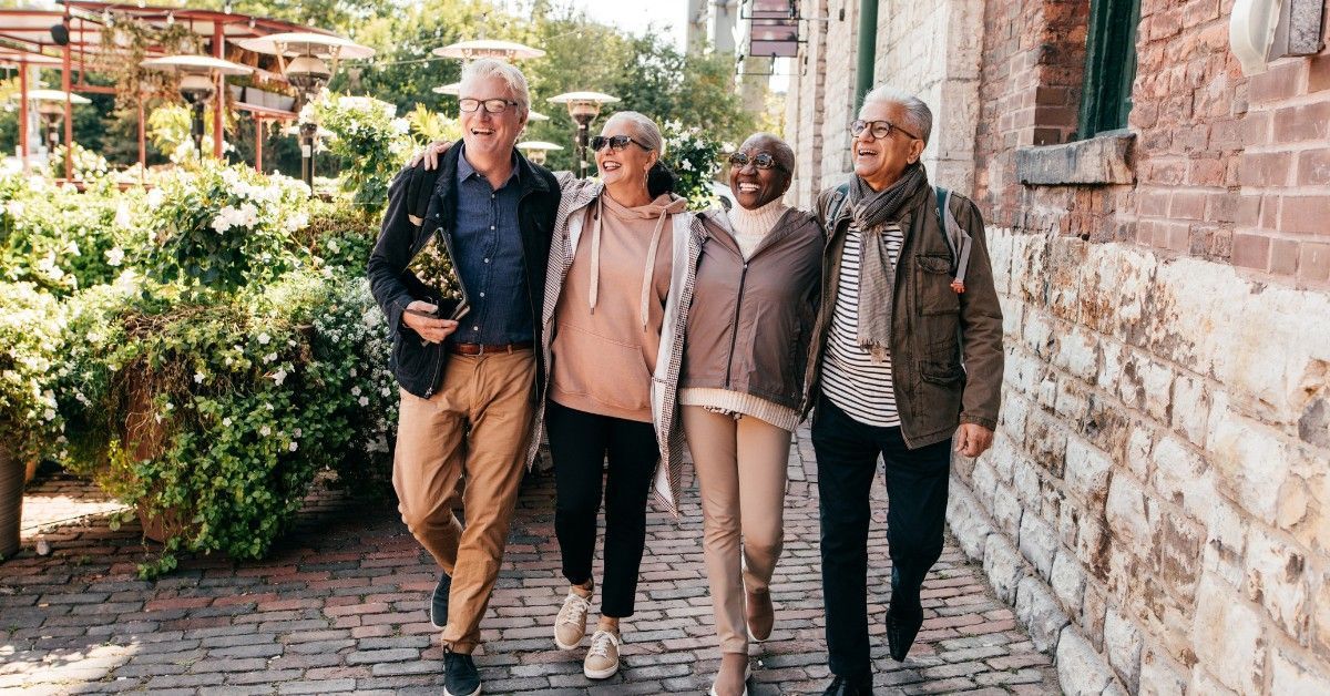 Four friends walking arm-in-arm along a brick path beside a wall and garden, smiling outdoors.