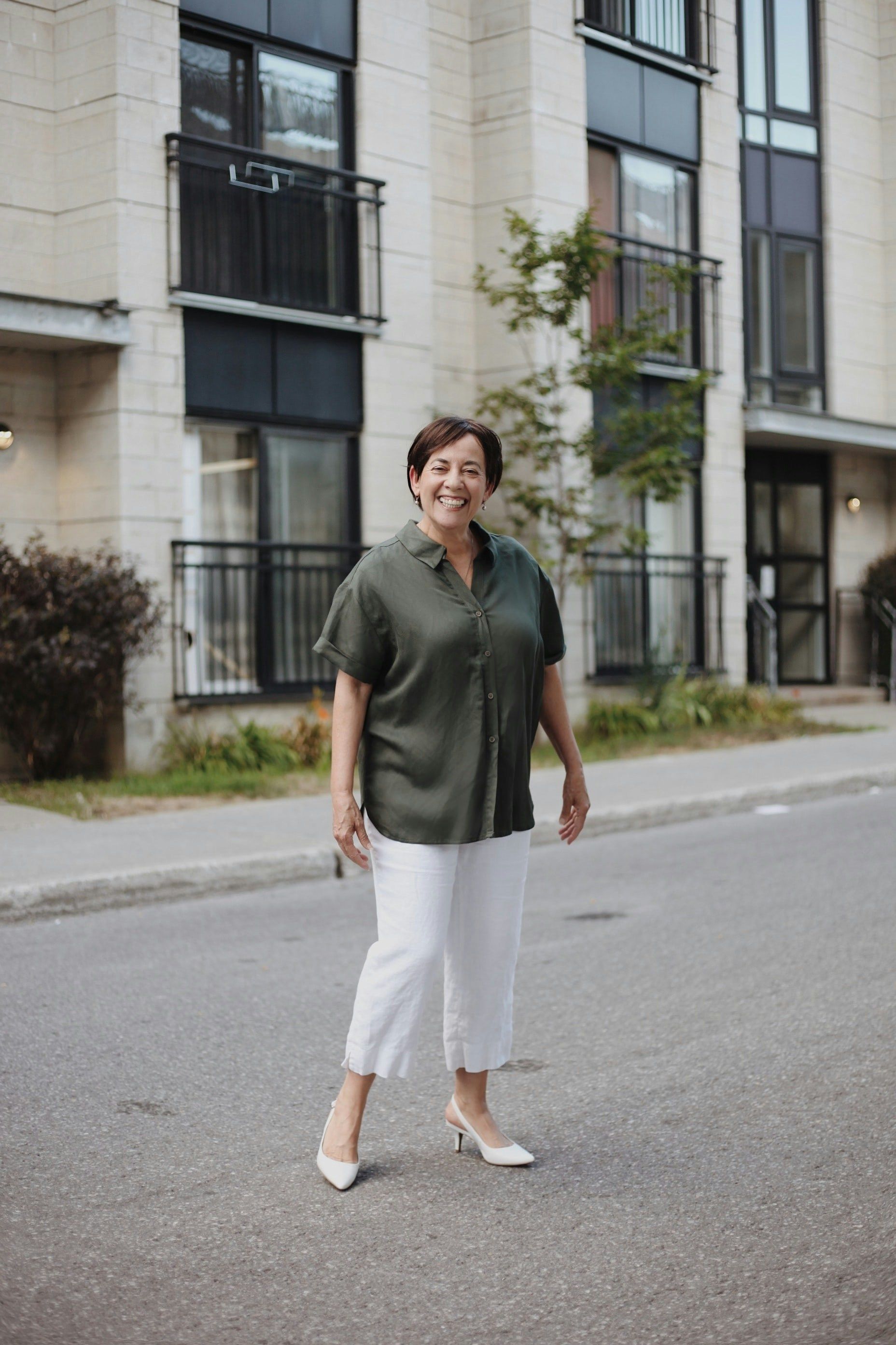 Woman standing on a street in front of apartment buildings, wearing a green shirt and white pants.