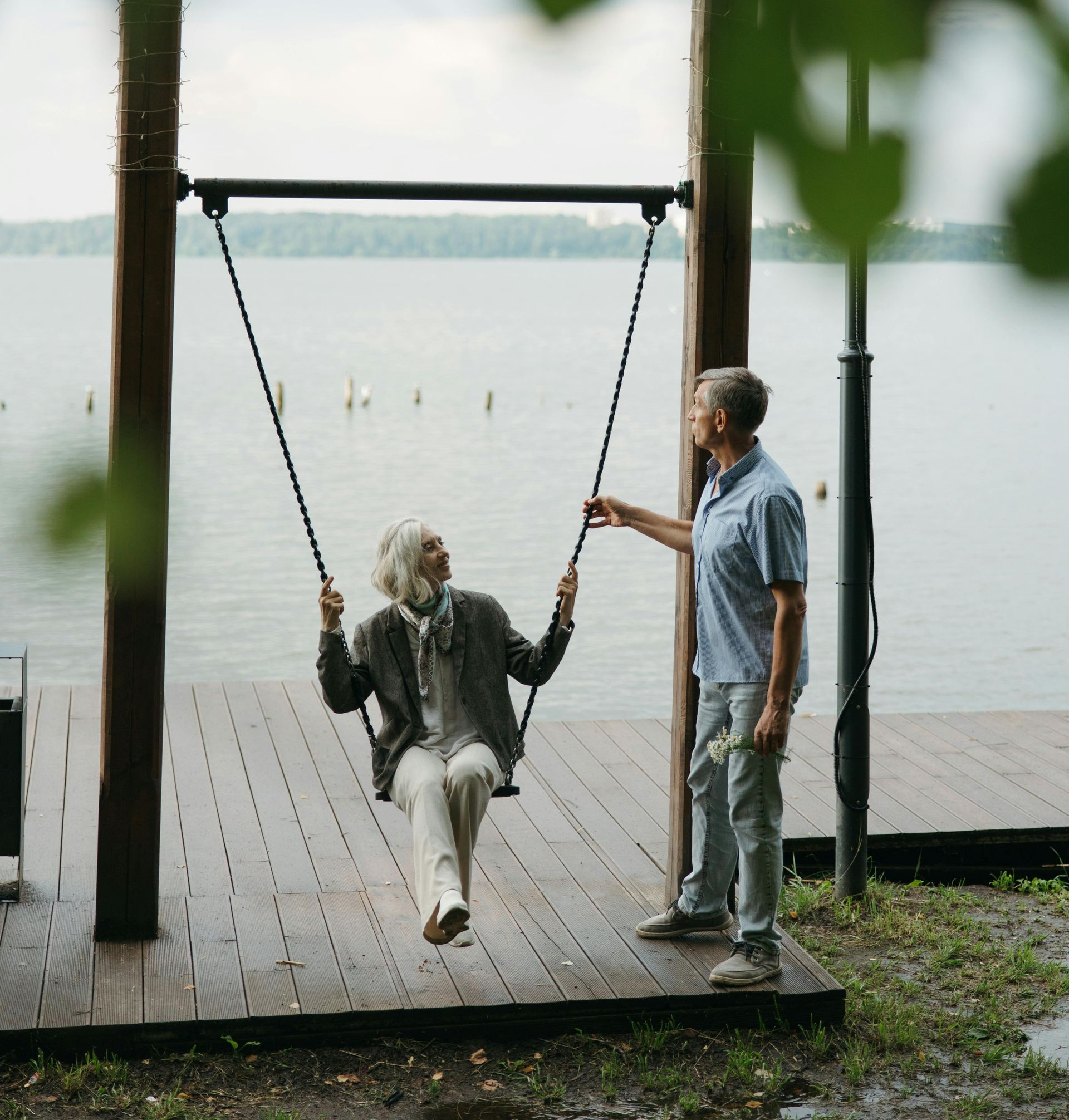 Two people by a lakeside swing, one seated and one standing, with a calm water backdrop.