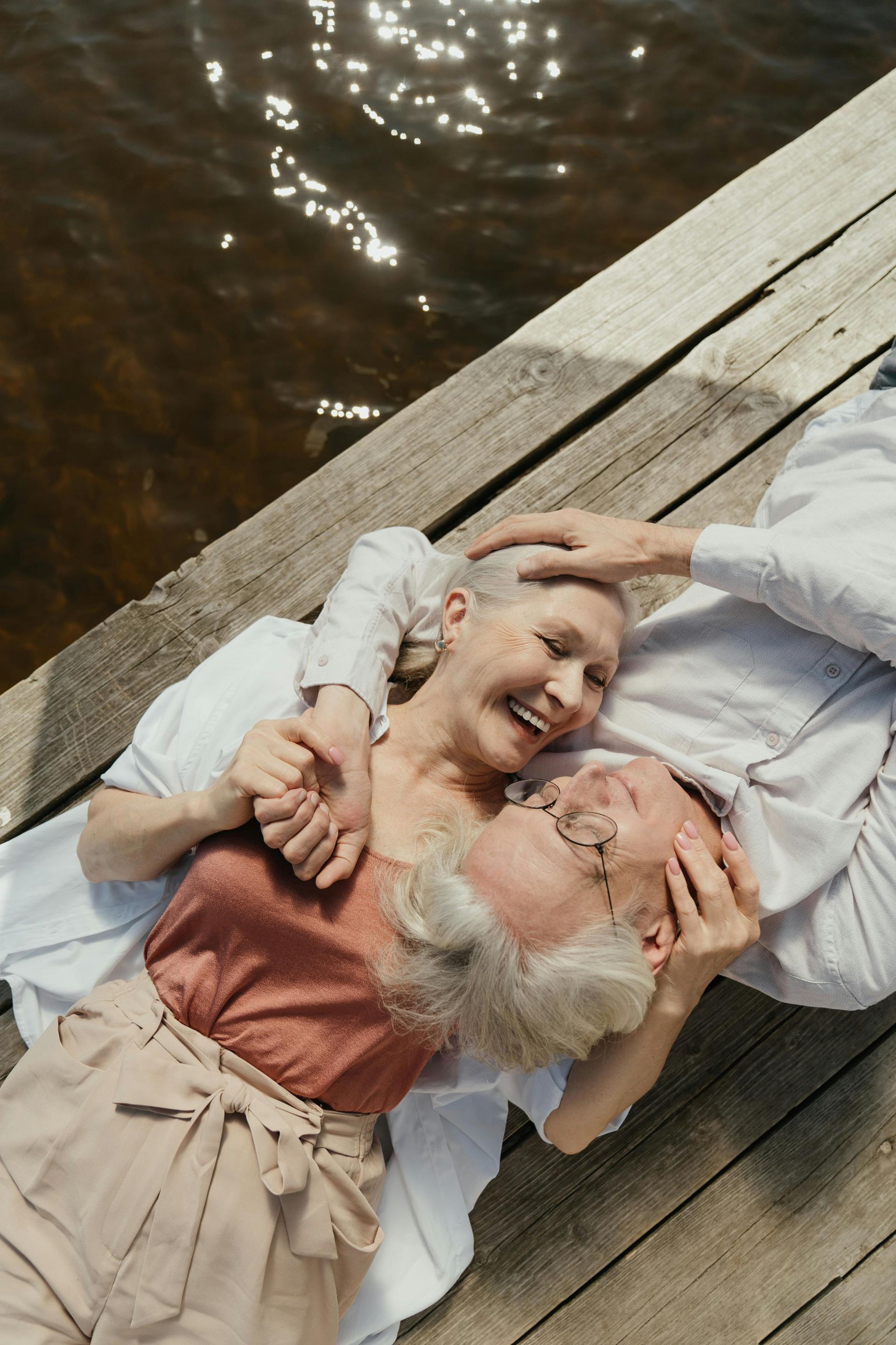 Two people lying on a dock, smiling and cuddling near the water.