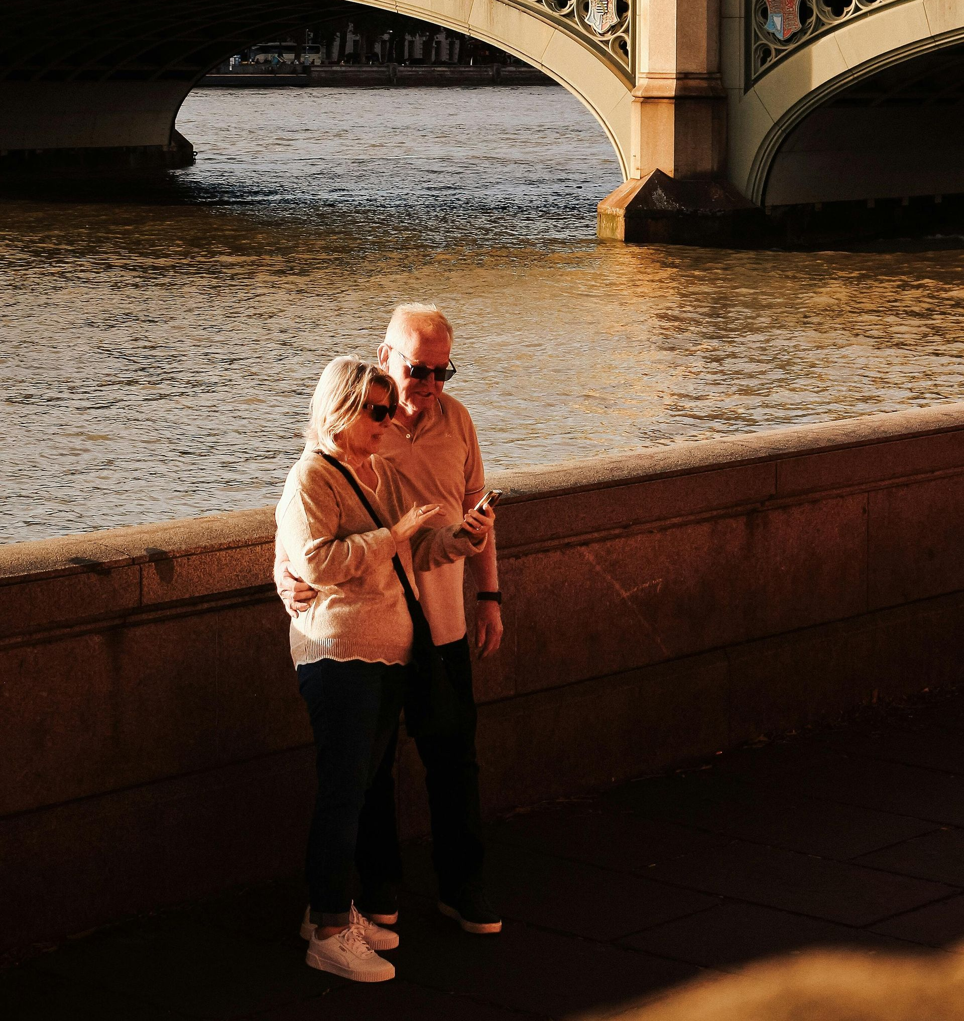 Two people standing by a riverside bridge, looking at a phone in warm evening light.