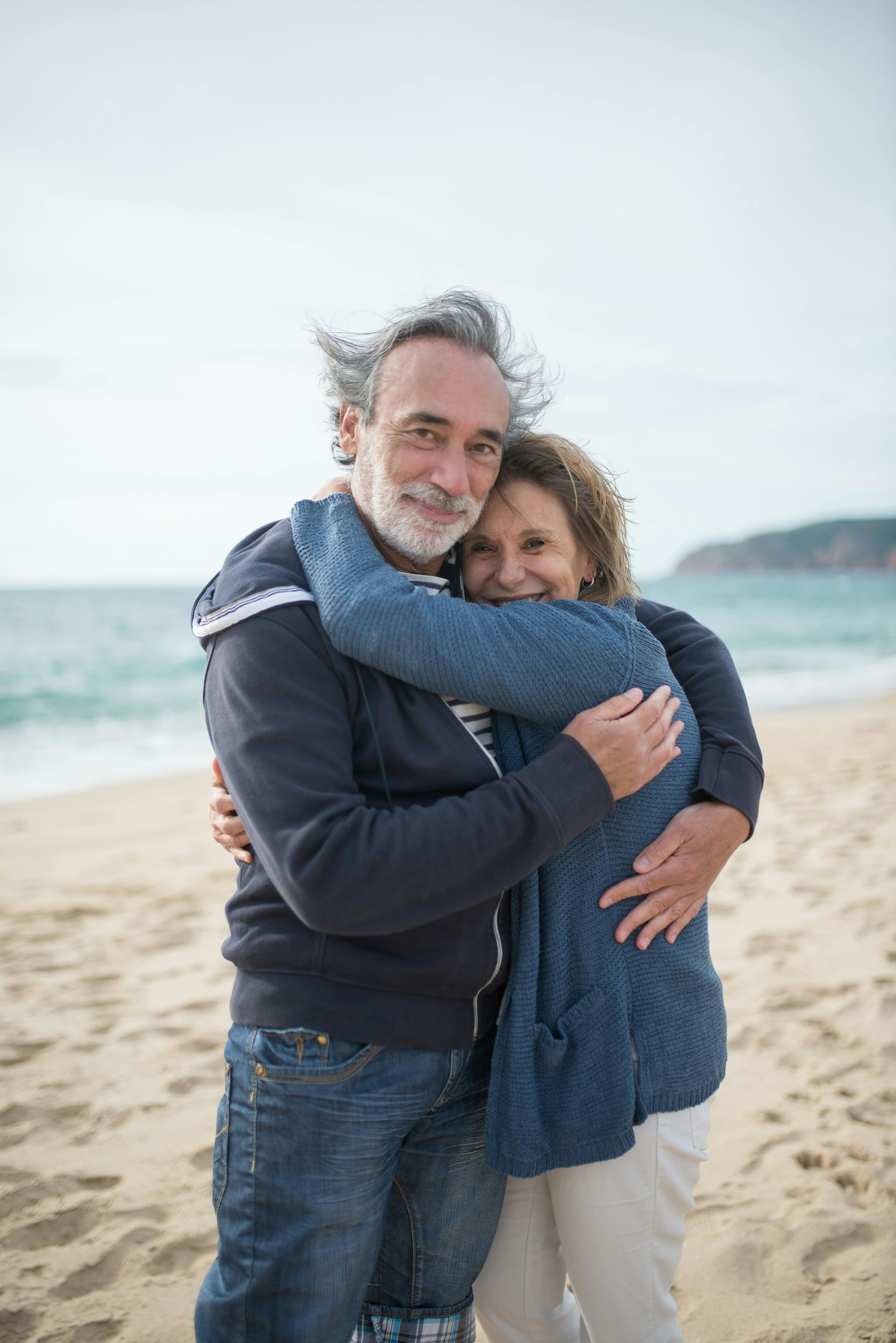 Couple hugging on a sandy beach, smiling and wrapped in blue sweaters.